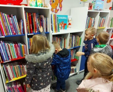 Na zdjęciu przedszkolaki przeglądają książki na półkach w bibliotece. - powiększ