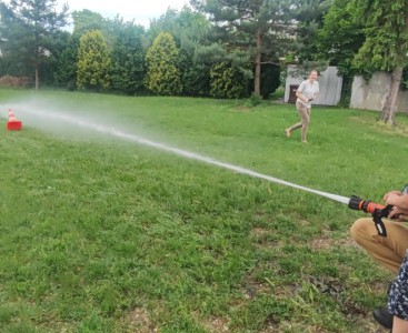 Zdjęcie przedstawia dziecko w trakcie lania wody z węża strażackiego - powiększ