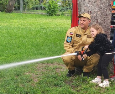 Zdjęcie przedstawia dziecko w trakcie lania wody z węża strażackiego - powiększ
