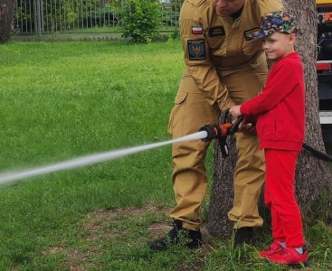 Zdjęcie przedstawia dziecko w trakcie lania wody z węża strażackiego - powiększ